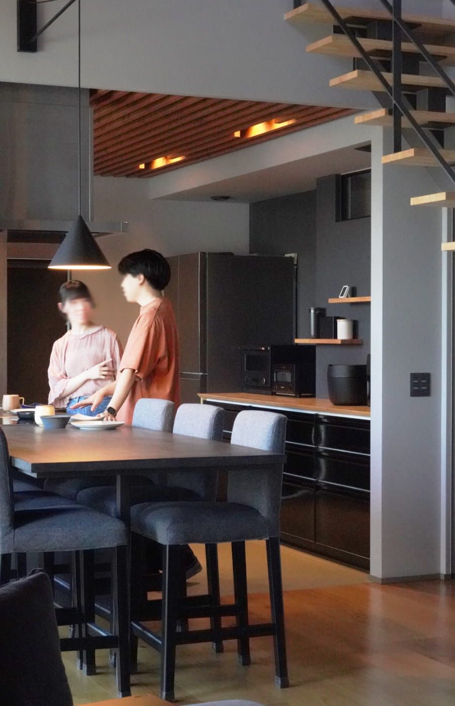 黒を基調としたモダンなキッチン。木の天井とアイアン階段が調和する洗練された空間で、二人がくつろいでいる様子。